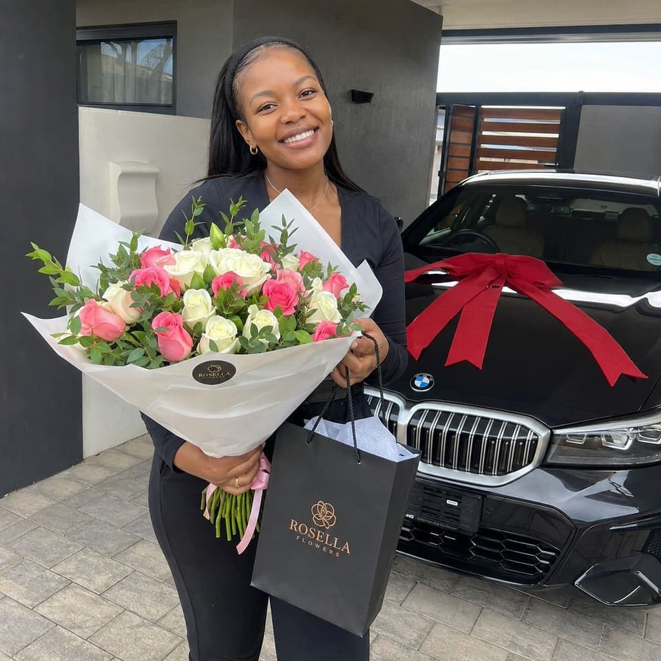 A graduate holding a pink and white bouquet beside a ribboned car