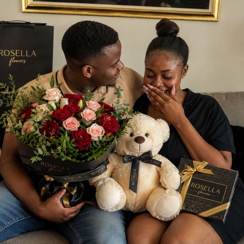 A couple sharing a large red rose arrangement, teddy bear and gifts